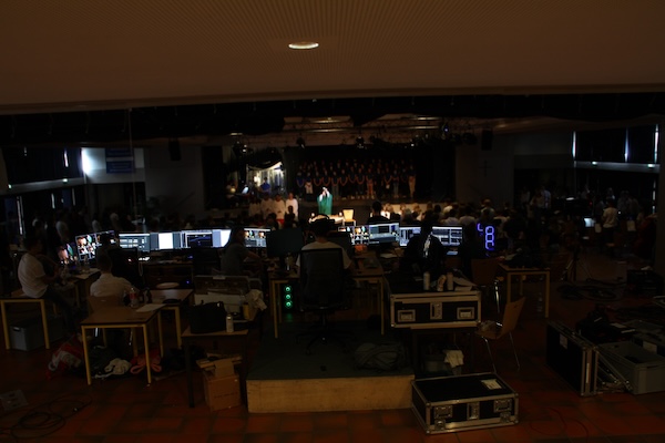Mischpulte, Monitore und Menschen von hinten, im Hintergrund eine BÃ¼hne mit Chor, bunt beleuchtet
